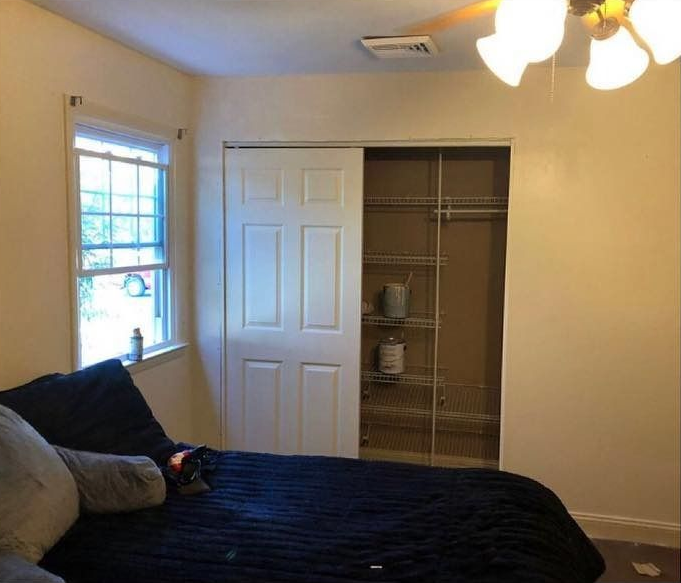 A bedroom with a dark blue bed, a white-framed window on the left, and a partially open closet with wire shelving.