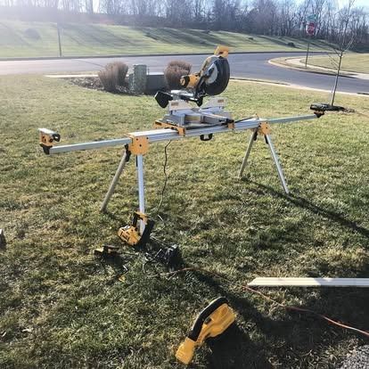 A yellow DeWalt miter saw mounted on a portable extension stand, set up on a grassy lawn near a road.