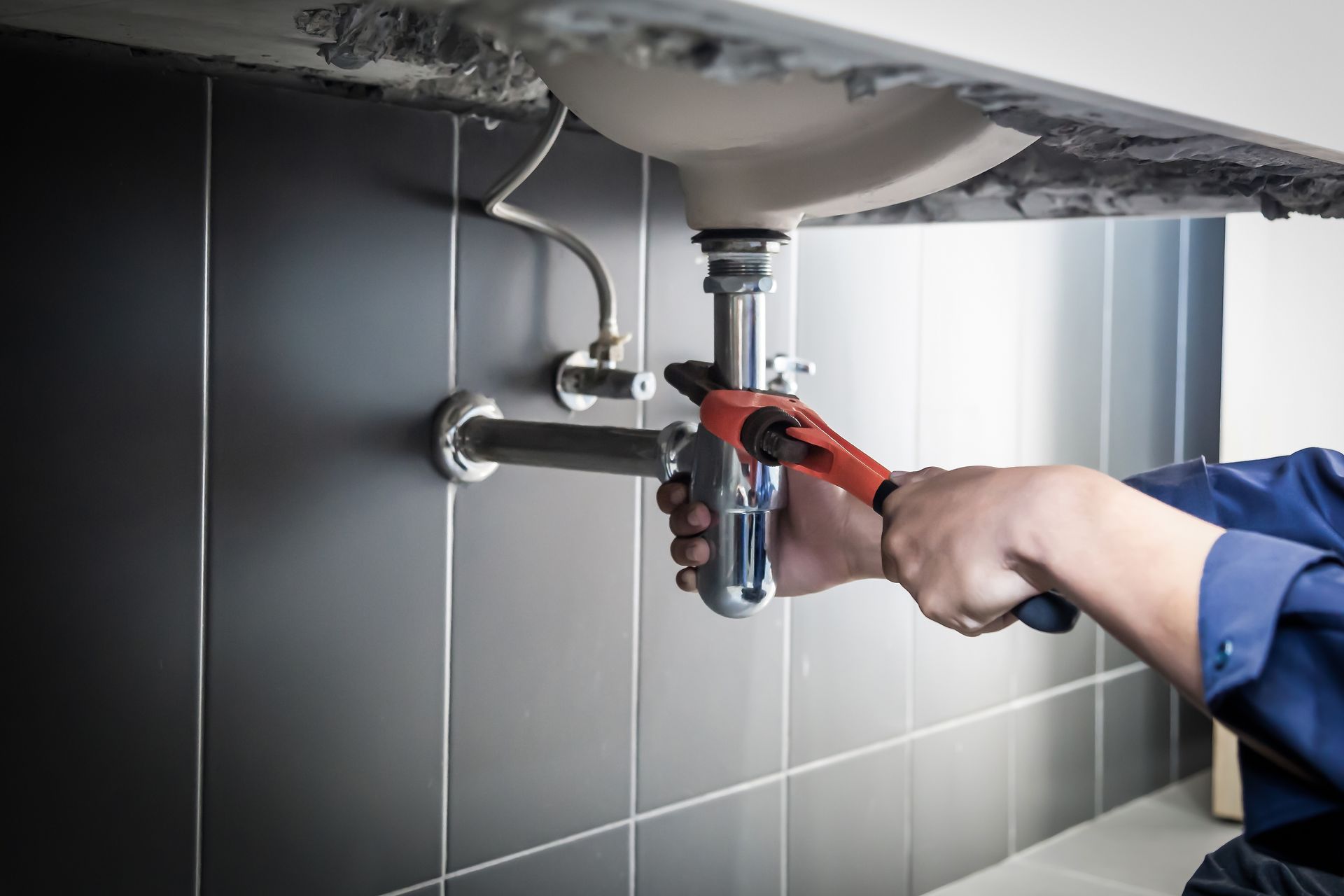 A person in a blue shirt uses a red pipe wrench to tighten a metal drain pipe under a sink against a gray tiled wall.