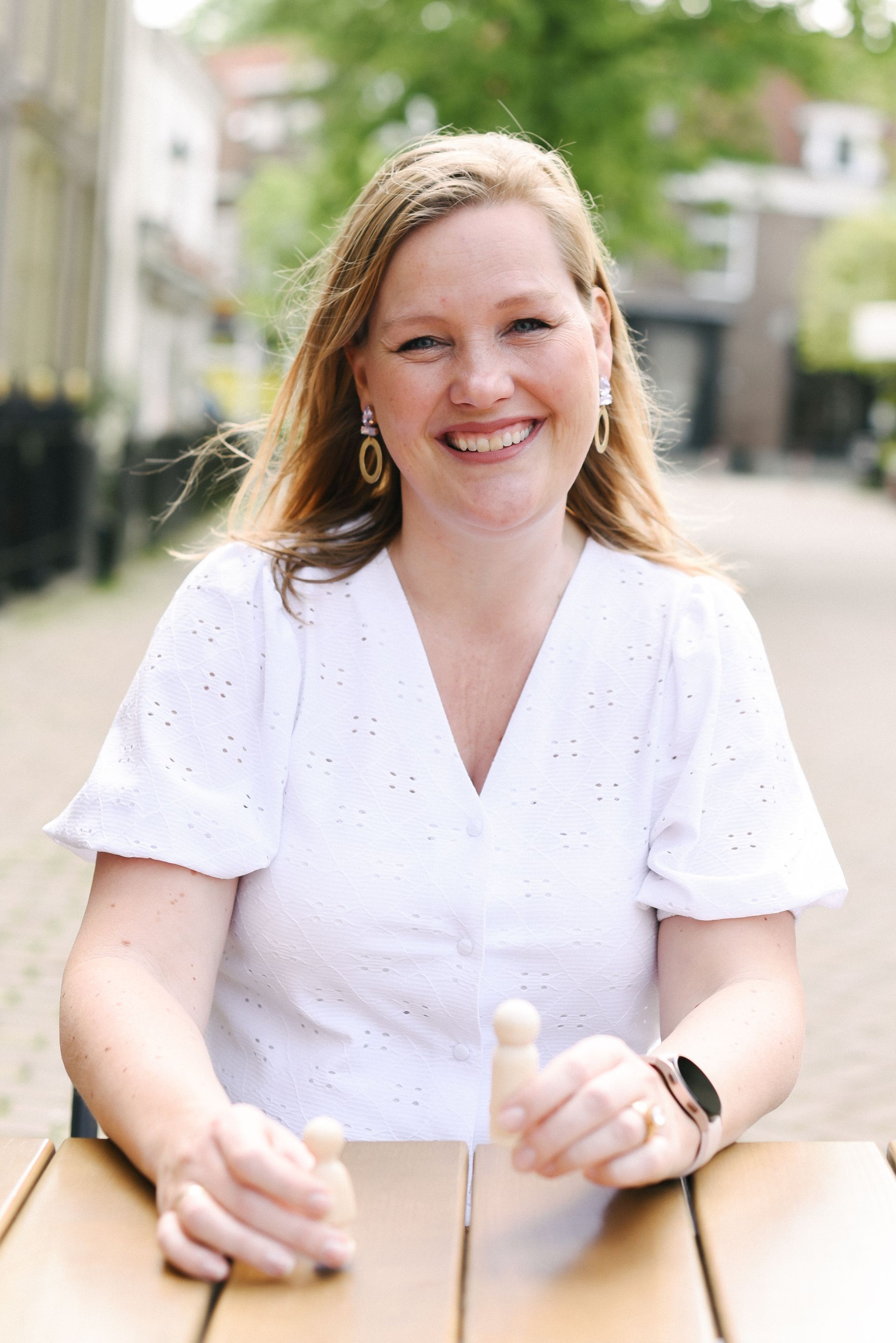 Een glimlachende persoon in een witte blouse met ajourwerk zit aan een buitentafel en houdt kleine houten figuurtjes in de handen.