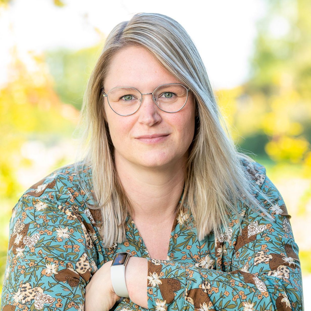 Een persoon met schouderlang blond haar en een bril met een dun metalen montuur draagt een groene blouse met bloemenprint in de buitenlucht.