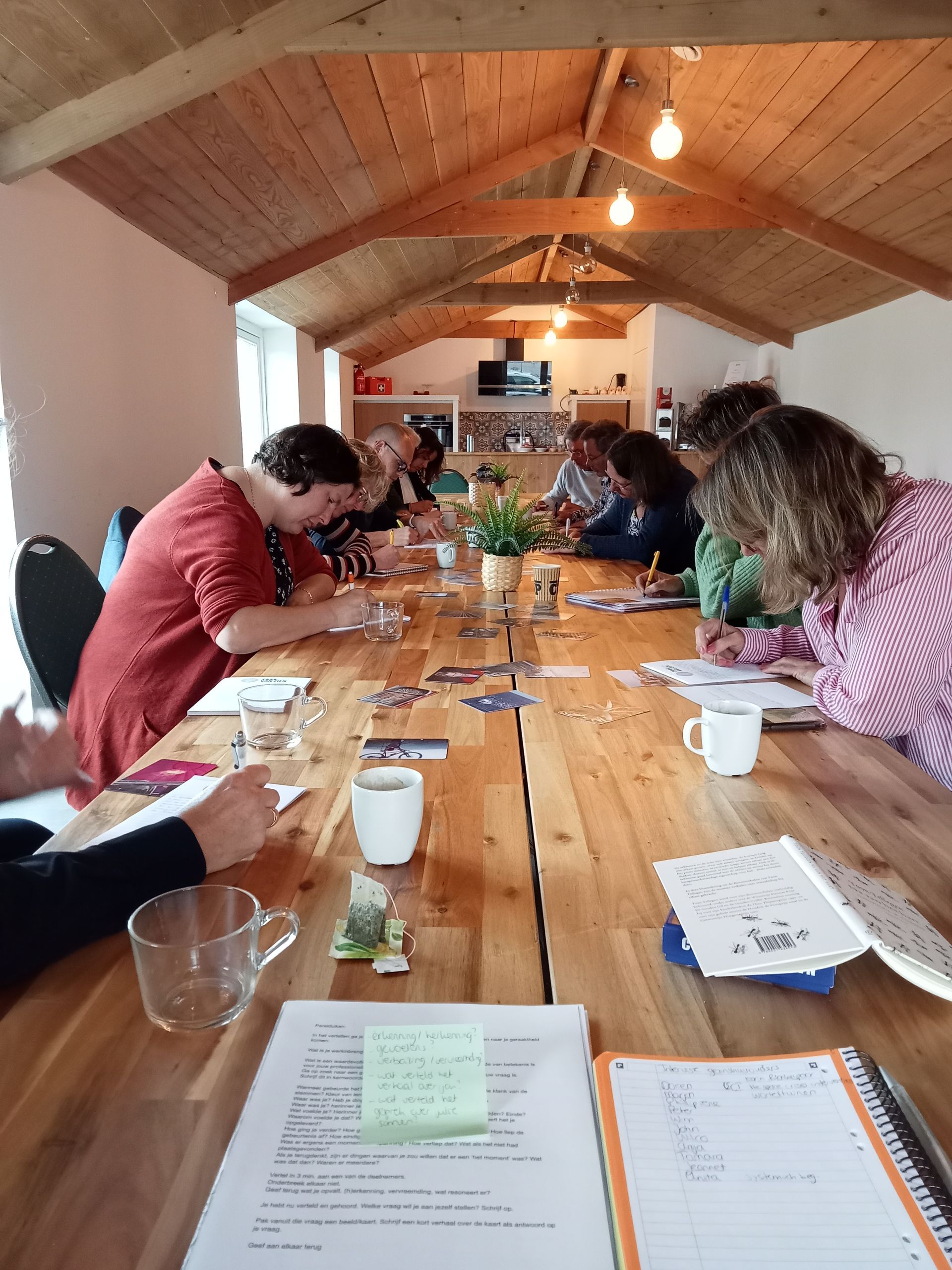 Een groep mensen zit rond een lange houten tafel in een rustieke kamer met een hoog plafond, bezig met het schrijven van documenten en het maken van aantekeningen.