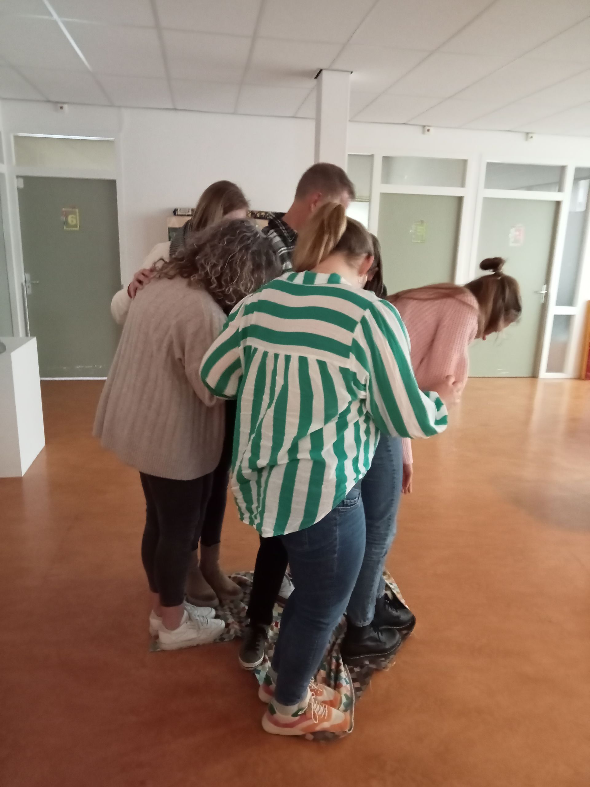 Vijf mensen staan ​​samen op een klein kleed in een felverlichte ruimte en doen mee aan een teambuildingsoefening.