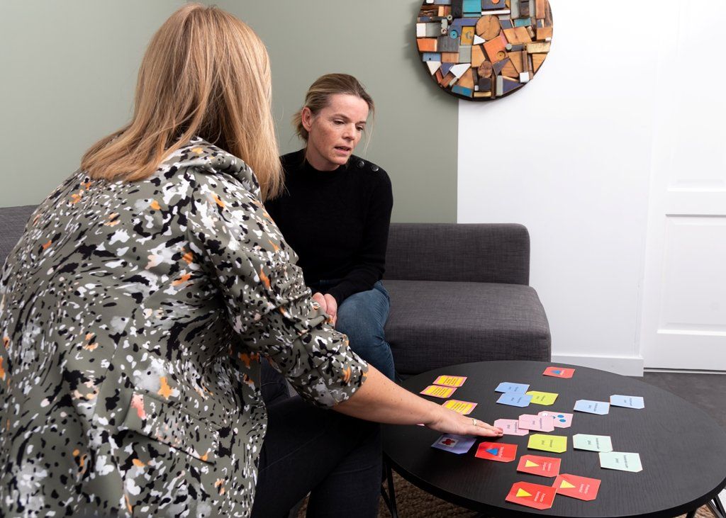 Twee personen zitten op een bank in een kantoor en bekijken gekleurde kaarten die uitgespreid liggen op een zwarte, ronde tafel.
