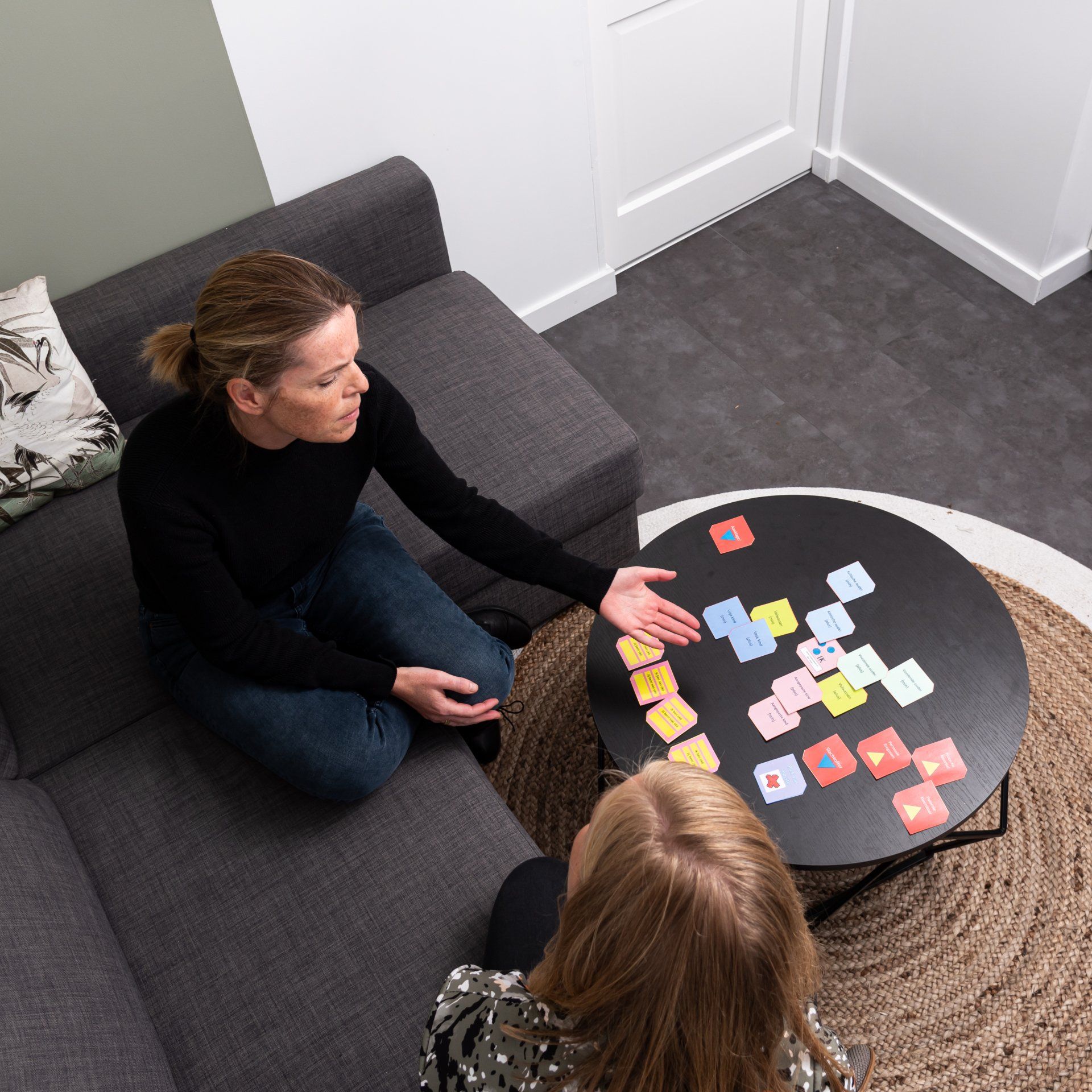 Twee mensen zitten op een bank en op de grond rond een tafel die bedekt is met kleurrijke notitiekaartjes en bespreken de rangschikking van de kaartjes.