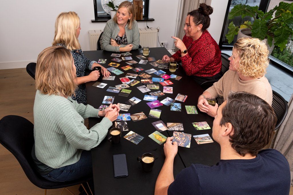 Zes mensen zitten rond een zwarte tafel en spelen een kaartspel met veel kaarten die over het tafelblad verspreid liggen.