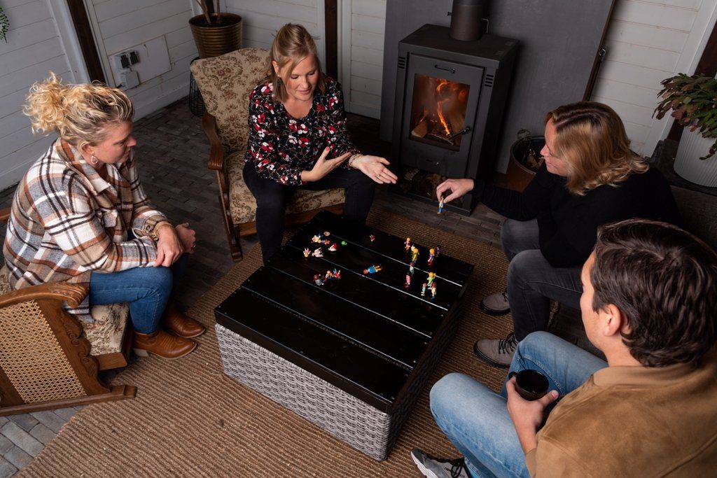 Vier mensen zitten op stoelen rond een tafel met spelstukken bij een brandende open haard in een gezellige binnenruimte.