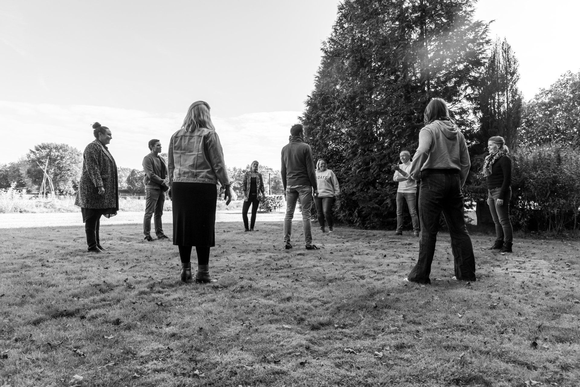 Een groep mensen staat in een kring op het gras in een park, van achteren gezien.