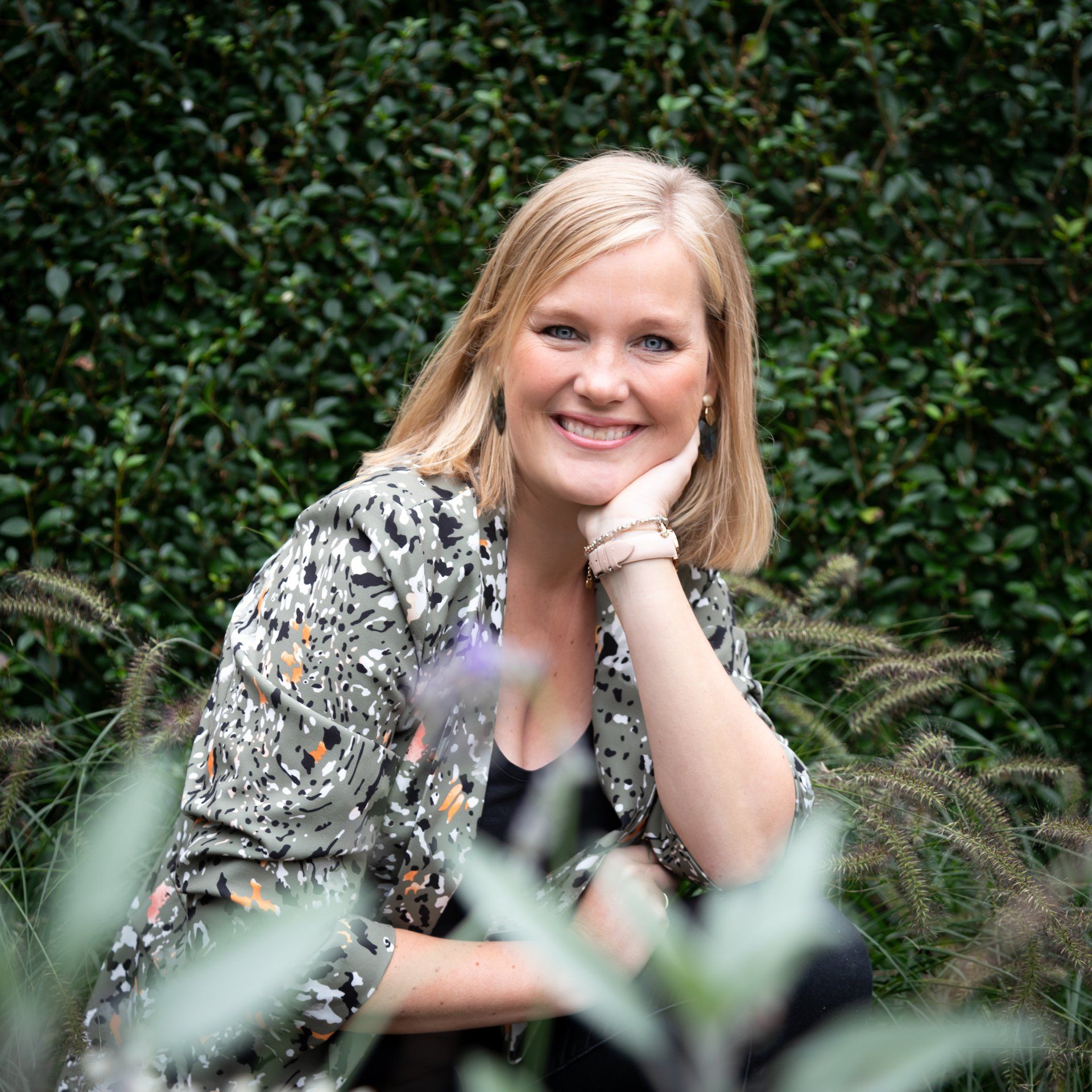Een persoon met blond haar glimlacht, met de kin op de hand, en draagt een jasje met een patroon in een tuin.