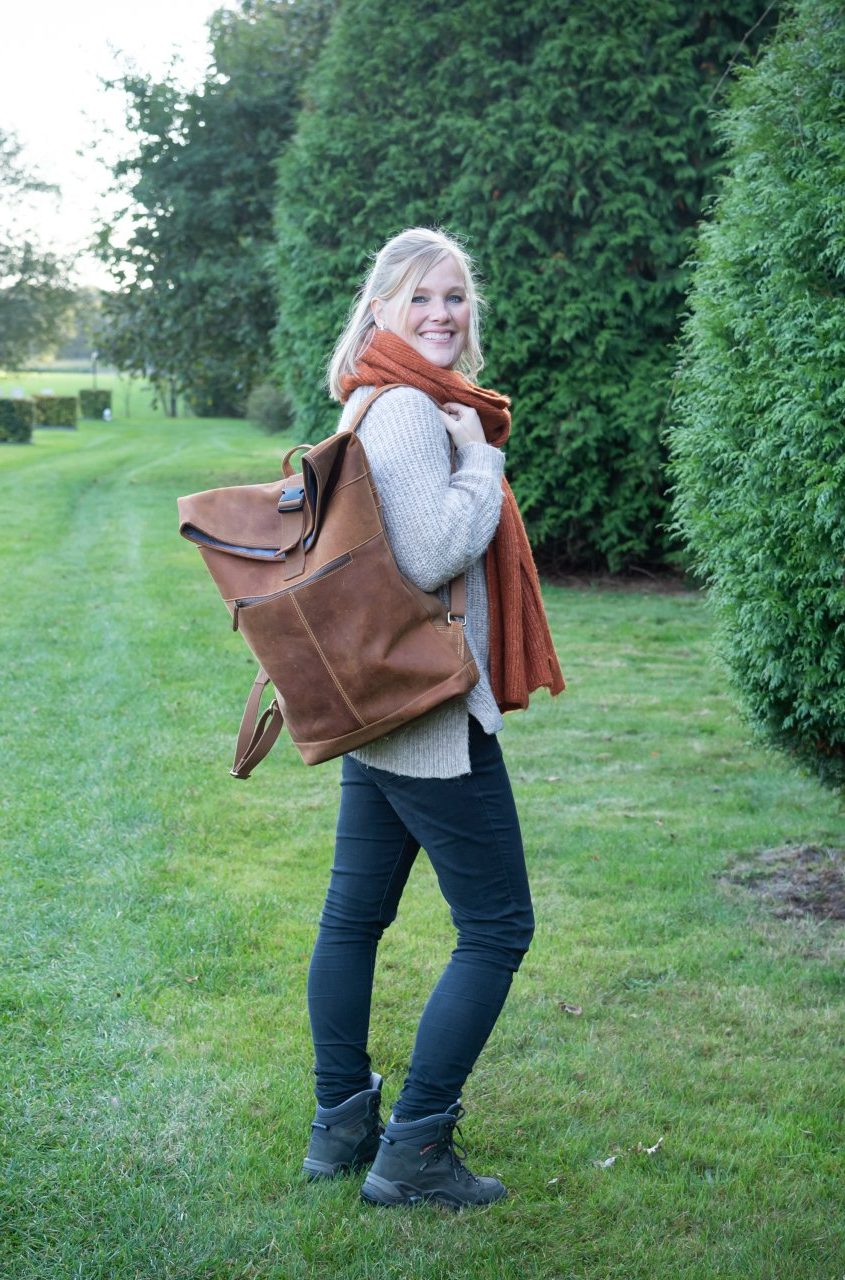 Een persoon lacht terwijl hij een bruine leren rugzak, een lichtgekleurde trui, een donkere broek en een oranje sjaal draagt, in een grasveld.