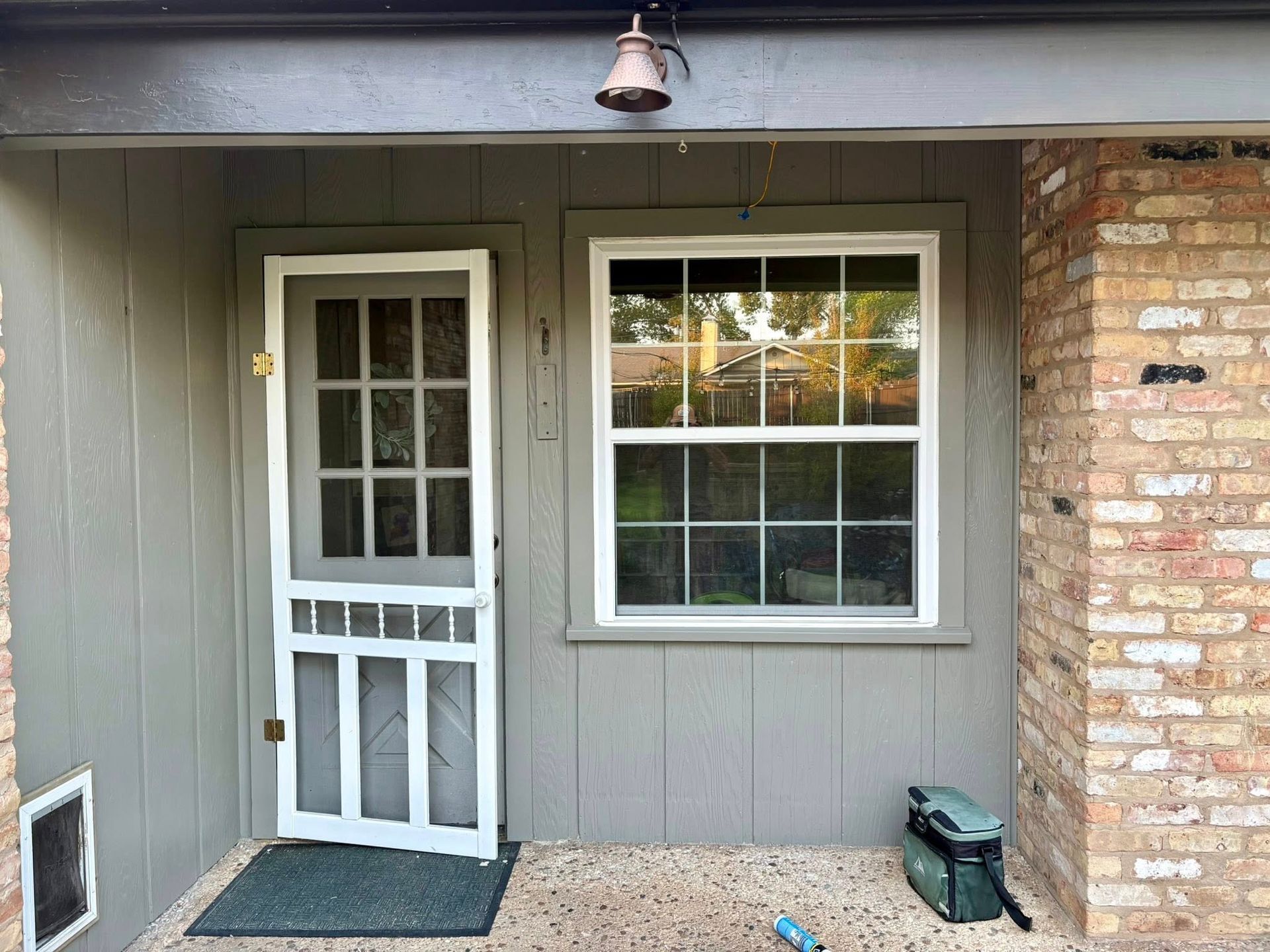 Front porch with gray siding, white door and window, brick wall, and a small green planter.