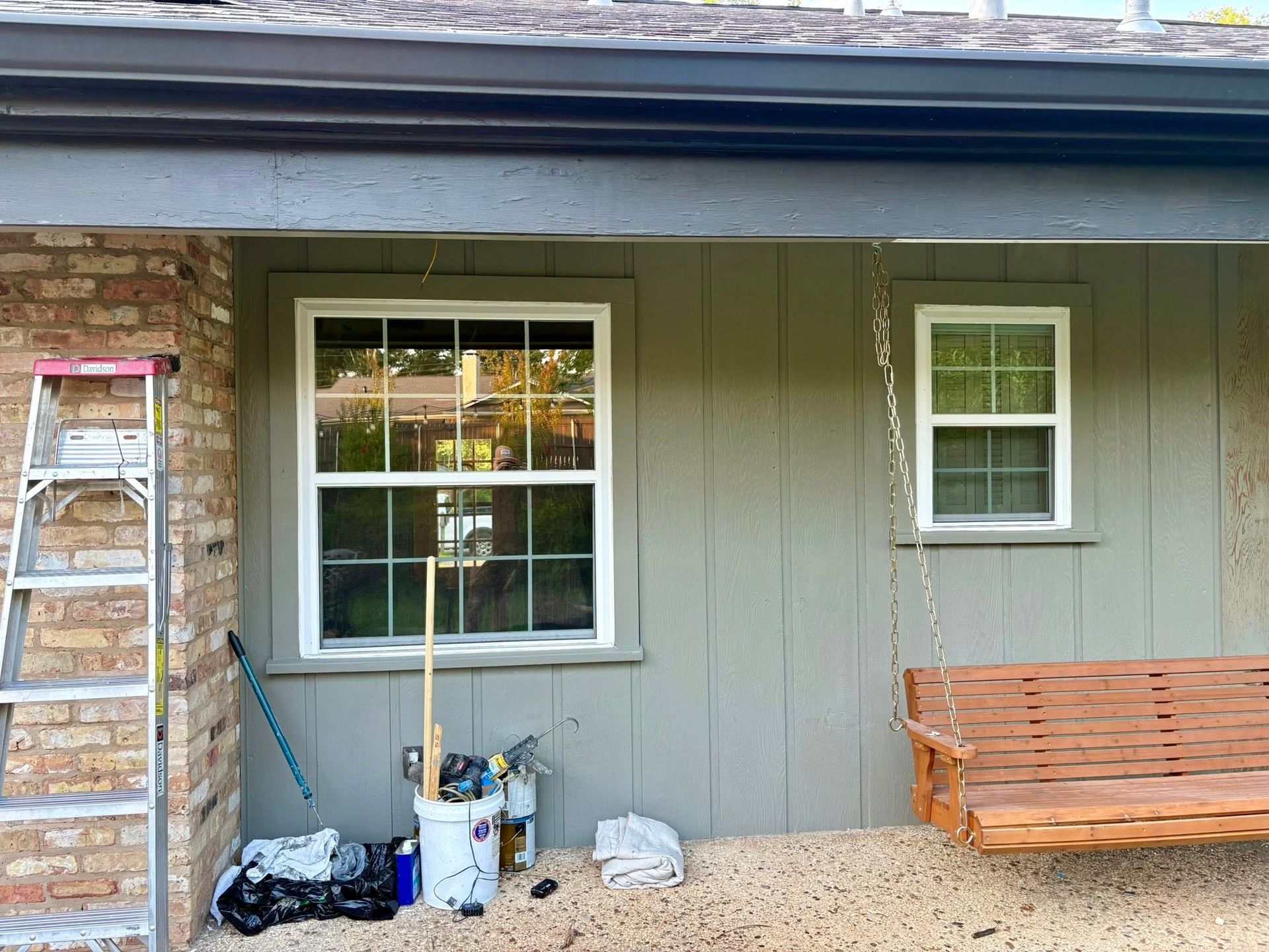 House exterior with gray siding, window, porch tools, and a wooden bench under the eaves