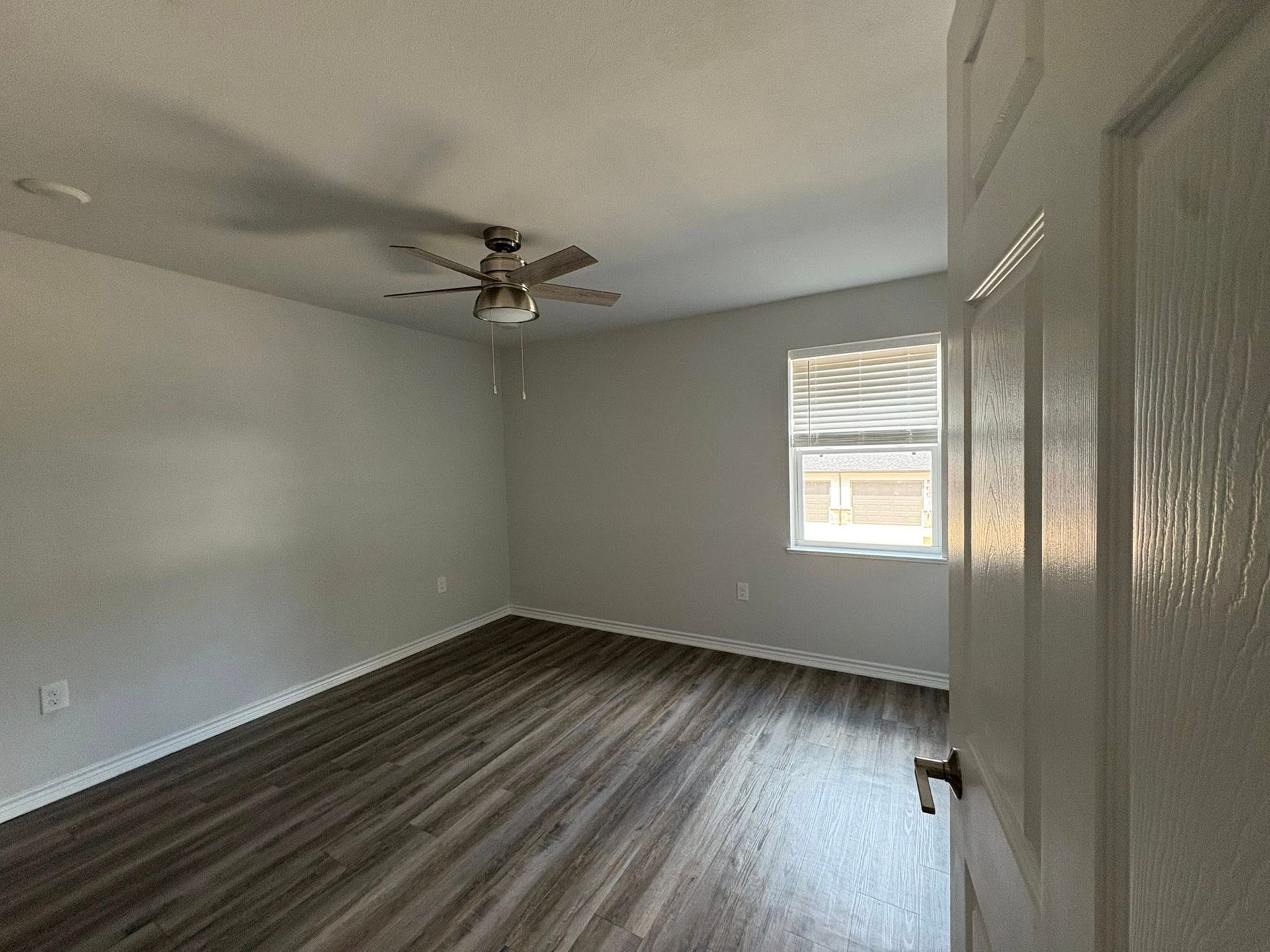 Empty gray bedroom with wood-look flooring, ceiling fan, and a window letting in daylight