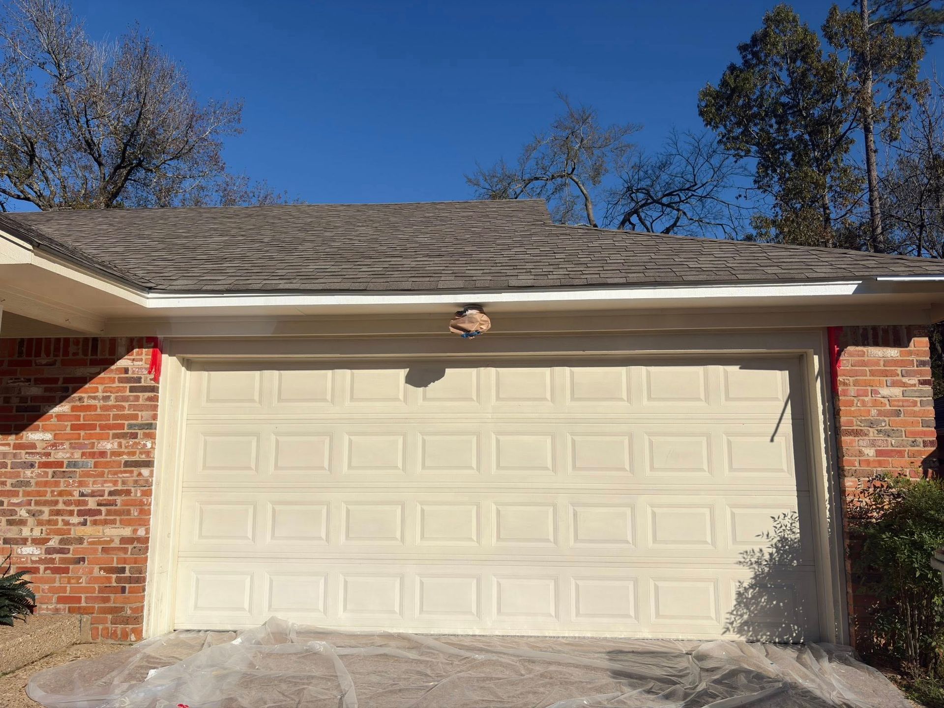 Residential garage with cream-colored door under a gray shingled roof, framed by red brick walls.