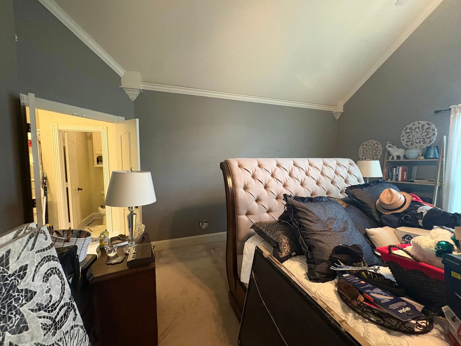 Bedroom with gray walls, tufted bed, nightstand lamp, and clothes scattered across the bed.