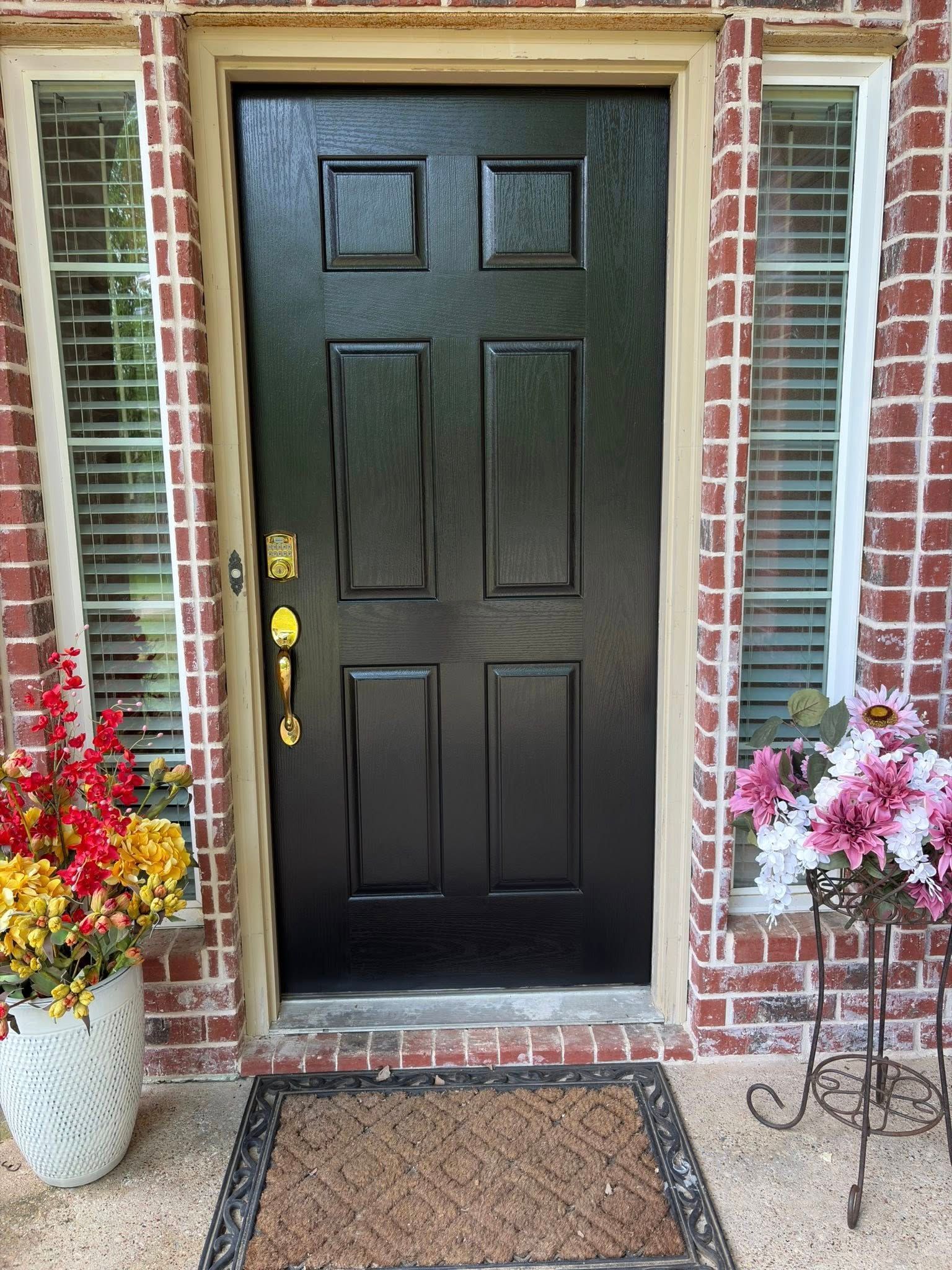 Front door with black paneling, brick trim, and potted flowers on both sides