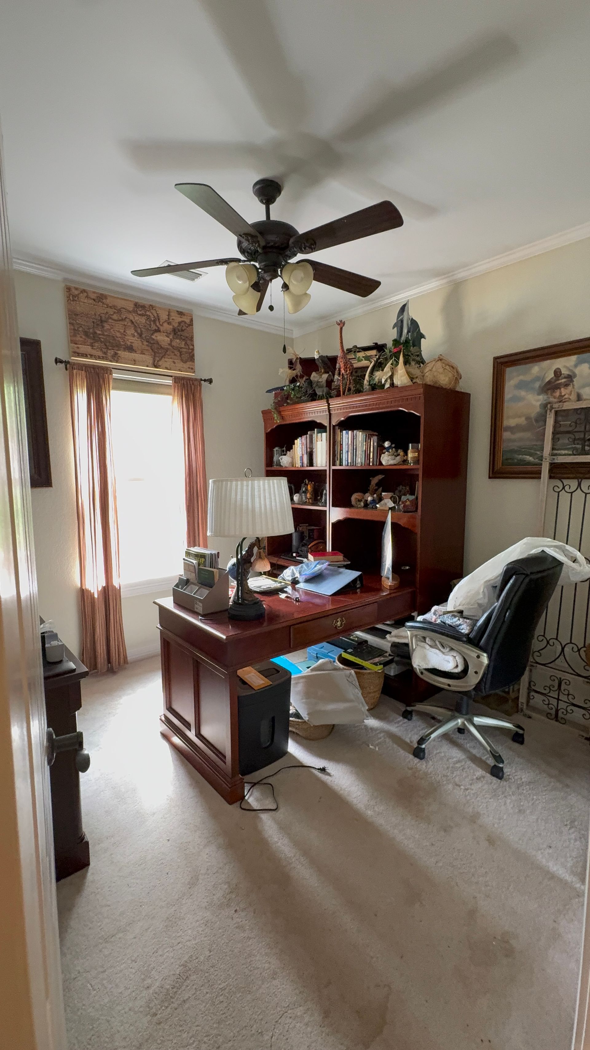 Home office with a wooden desk, black chair, ceiling fan, bookshelves, and a window with curtains.