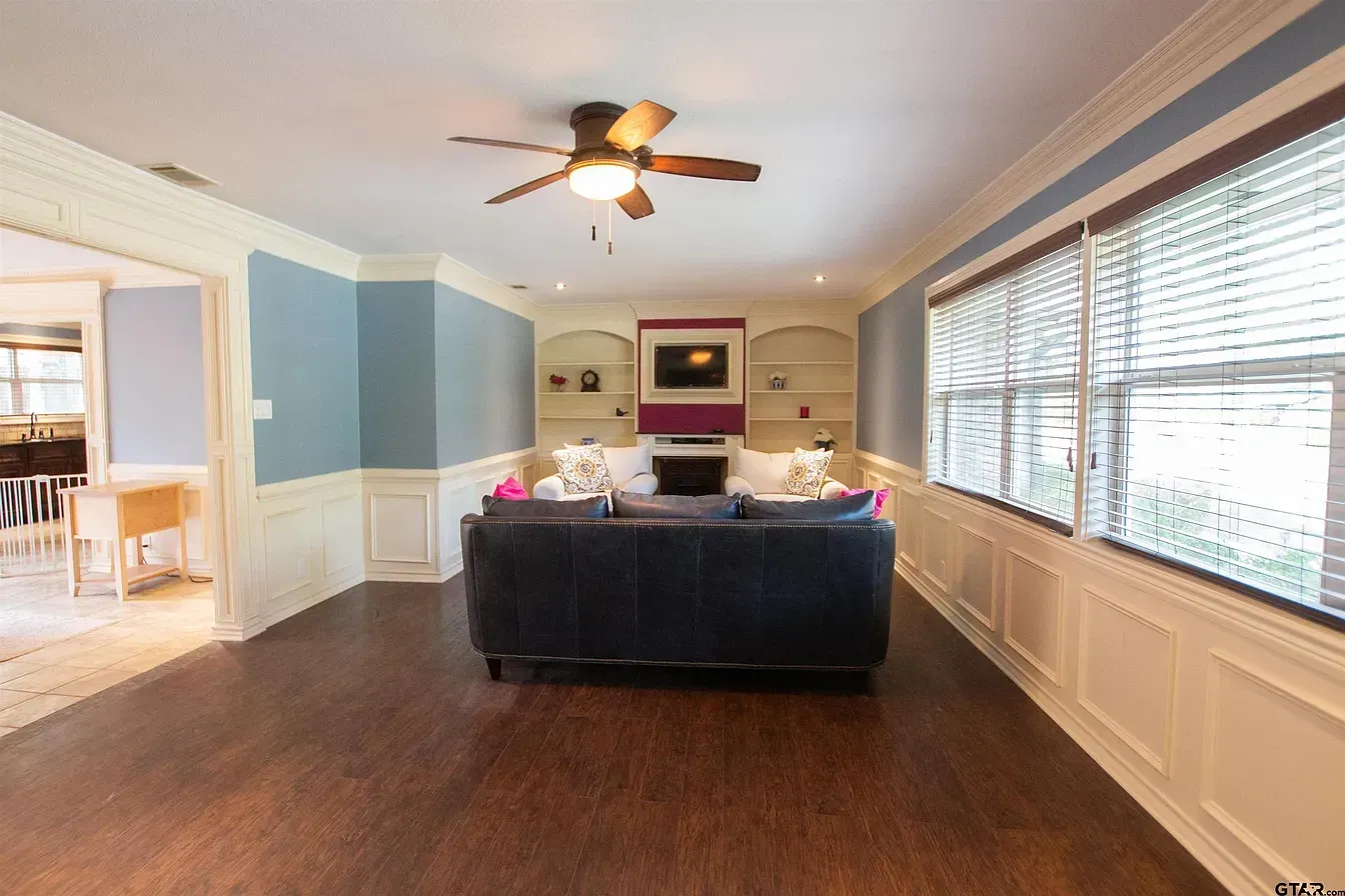 Spacious living room with hardwood floors, blue walls, a dark sofa, fireplace, ceiling fan, and large windows.