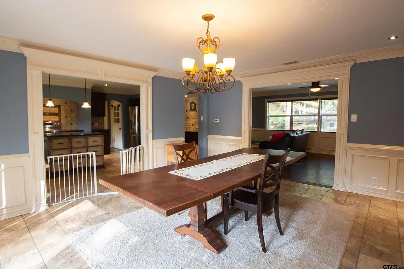 Dining room with wooden table, chandelier, blue walls, and tiled floor, opening to a kitchen and living area