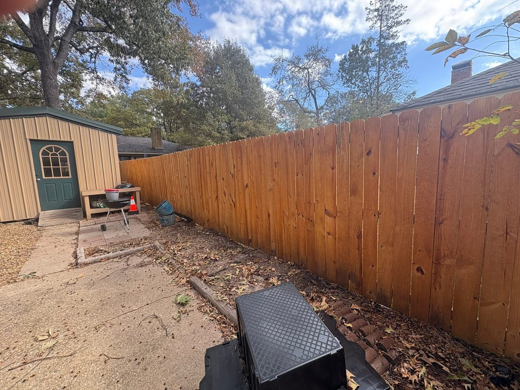 Backyard with wooden fence, gravel path, shed, and a black storage bin under a cloudy sky