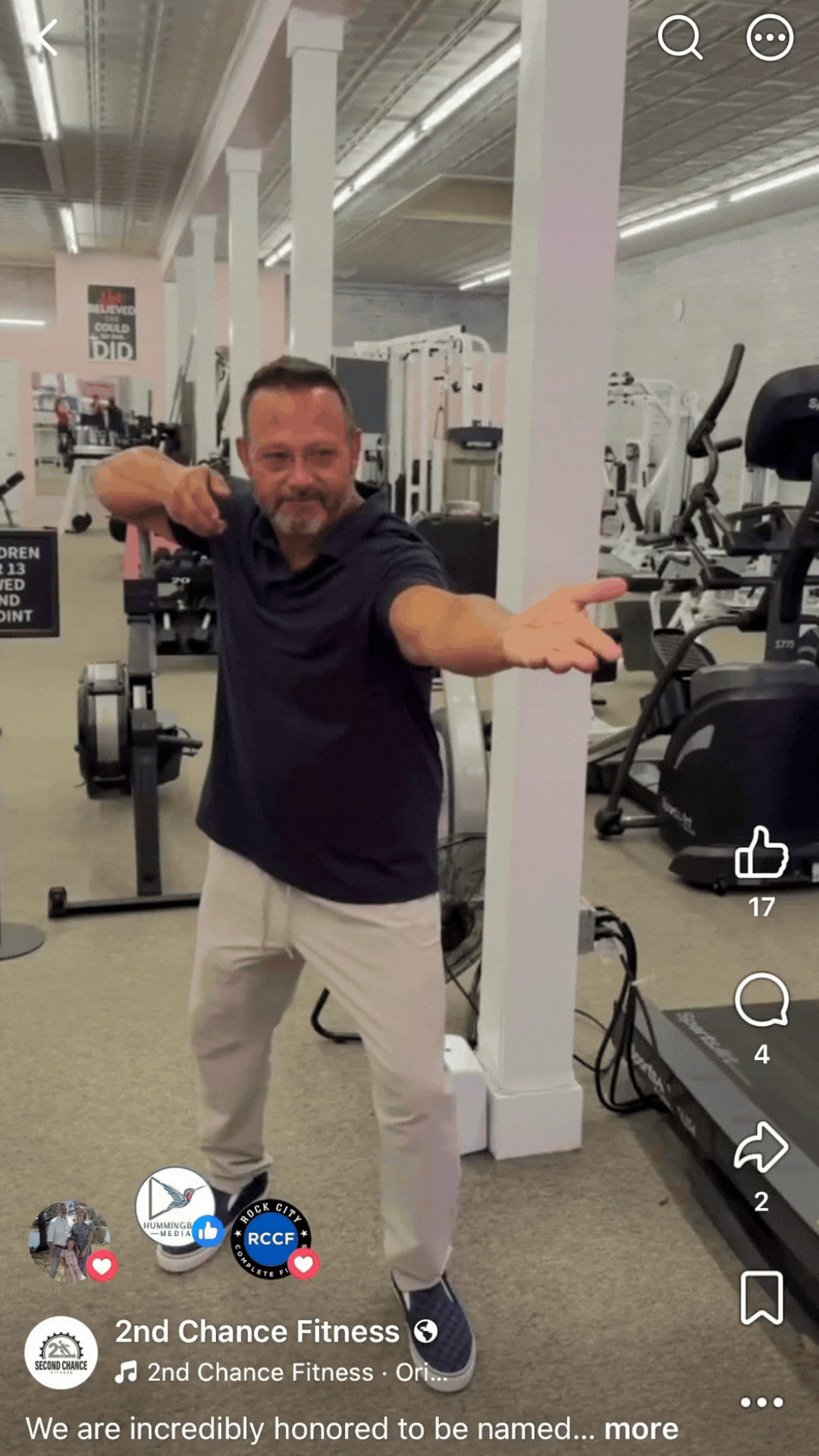 Man in dark shirt and light pants posing with outstretched arms in a gym.