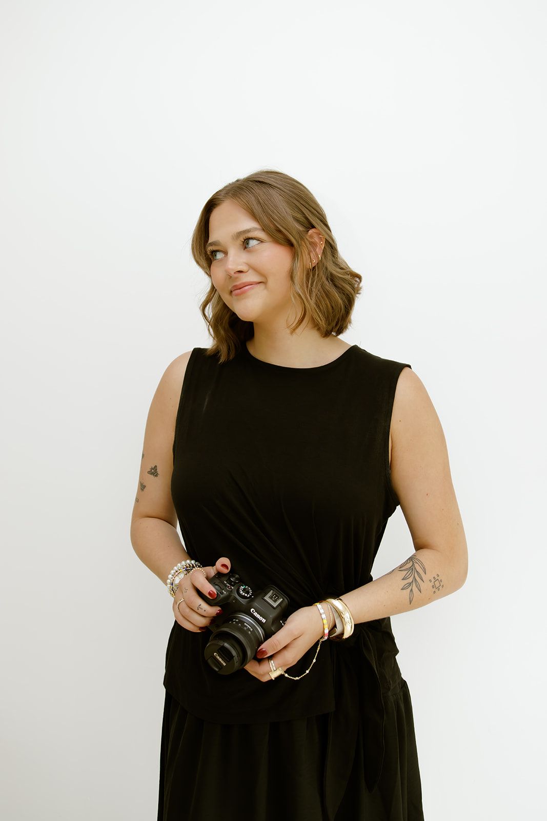 Woman in black outfit holding a camera, standing against a plain white background.