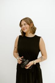 Woman in black outfit holding a camera, standing against a plain white background.