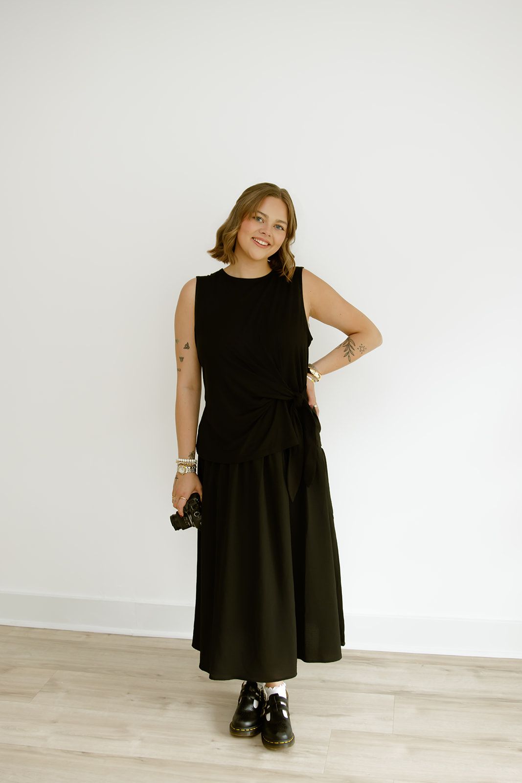 Woman in a black dress standing in a bright room, holding sunglasses and smiling.