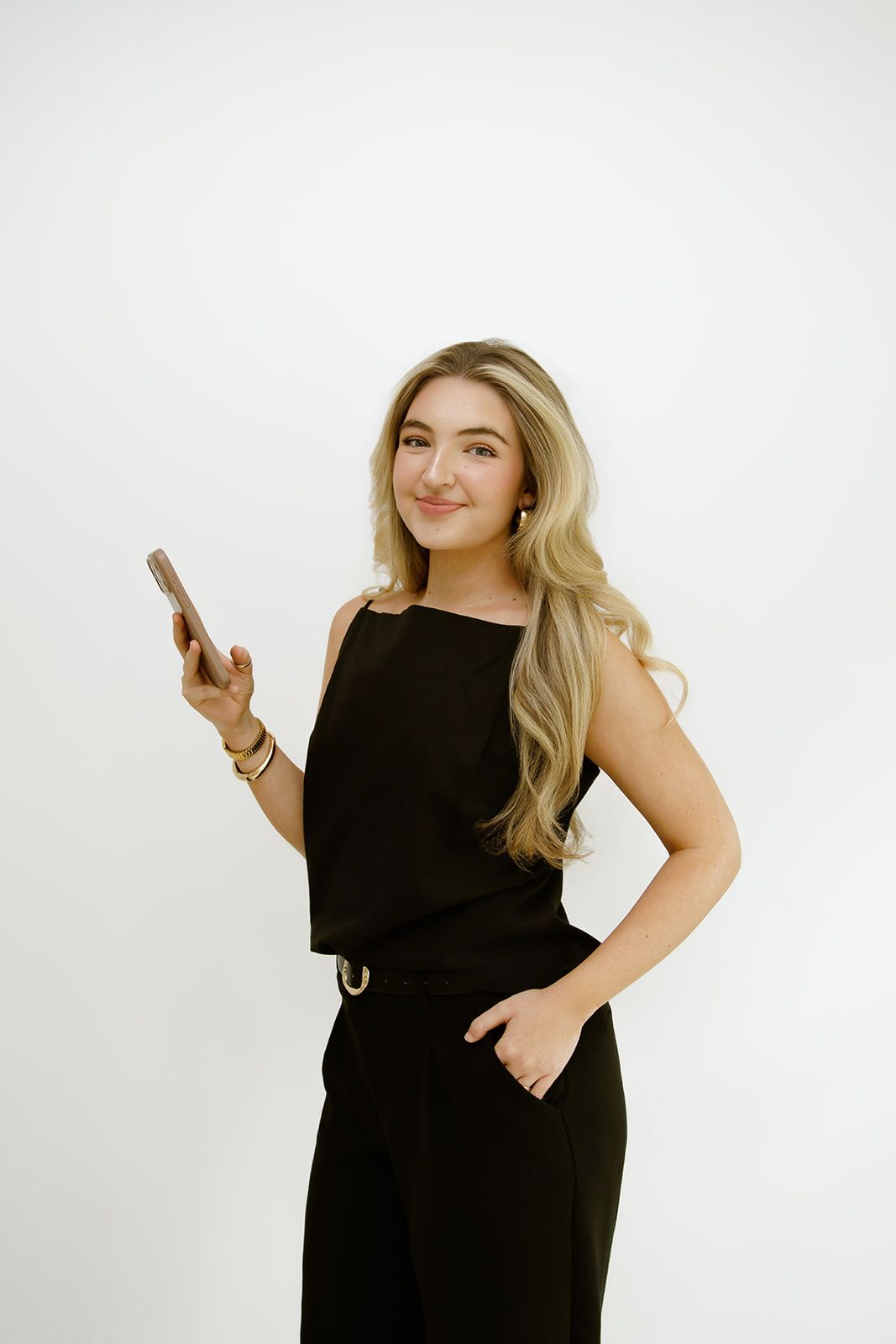 Woman in a black outfit holding a phone and smiling against a white background