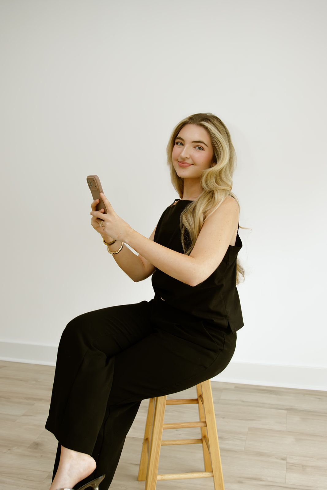 Person in black outfit sitting on a wooden stool, smiling and holding a phone against a plain white wall.
