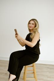 Person in black outfit sitting on a wooden stool, smiling and holding a phone against a plain white wall.