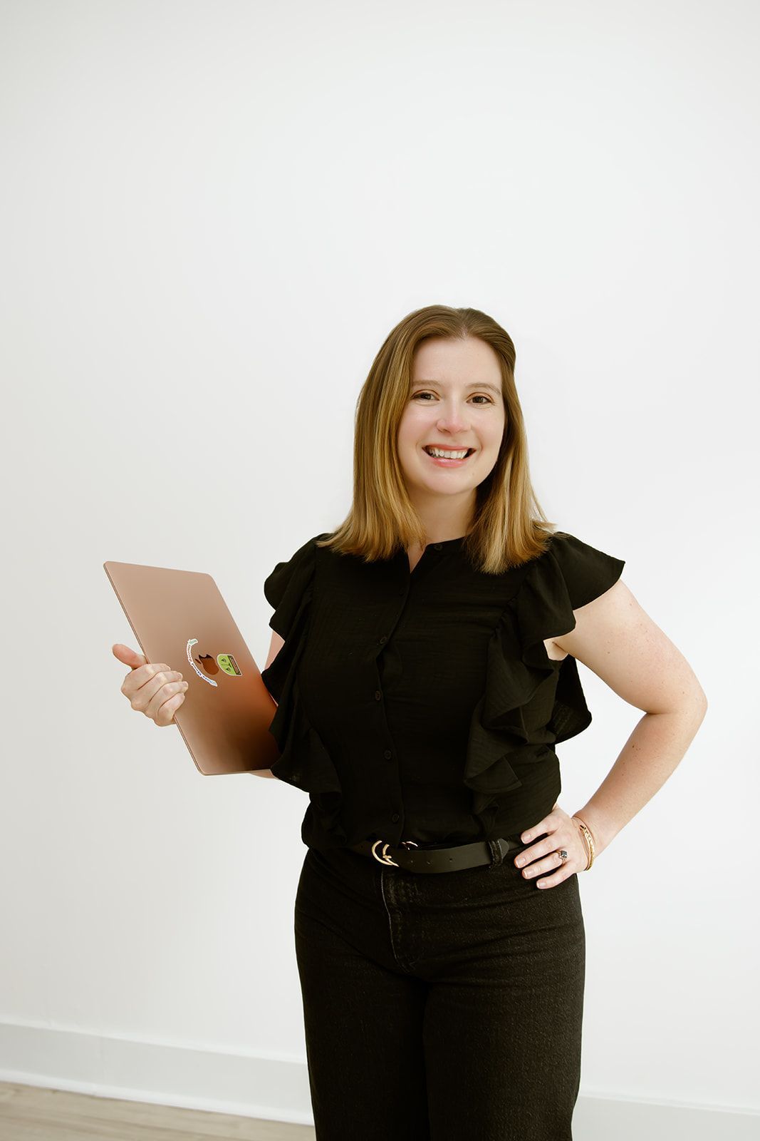 Smiling person in black outfit holding a clipboard, standing against a white wall.