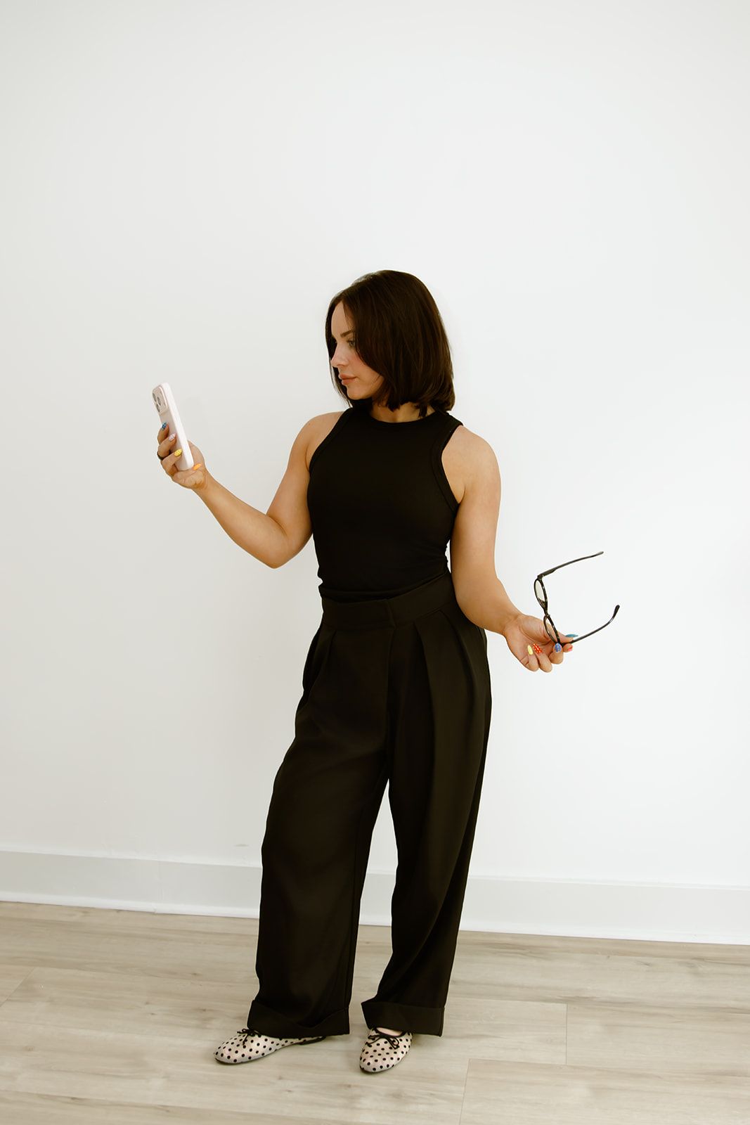 Woman in black outfit holding a phone and glasses against a white wall.