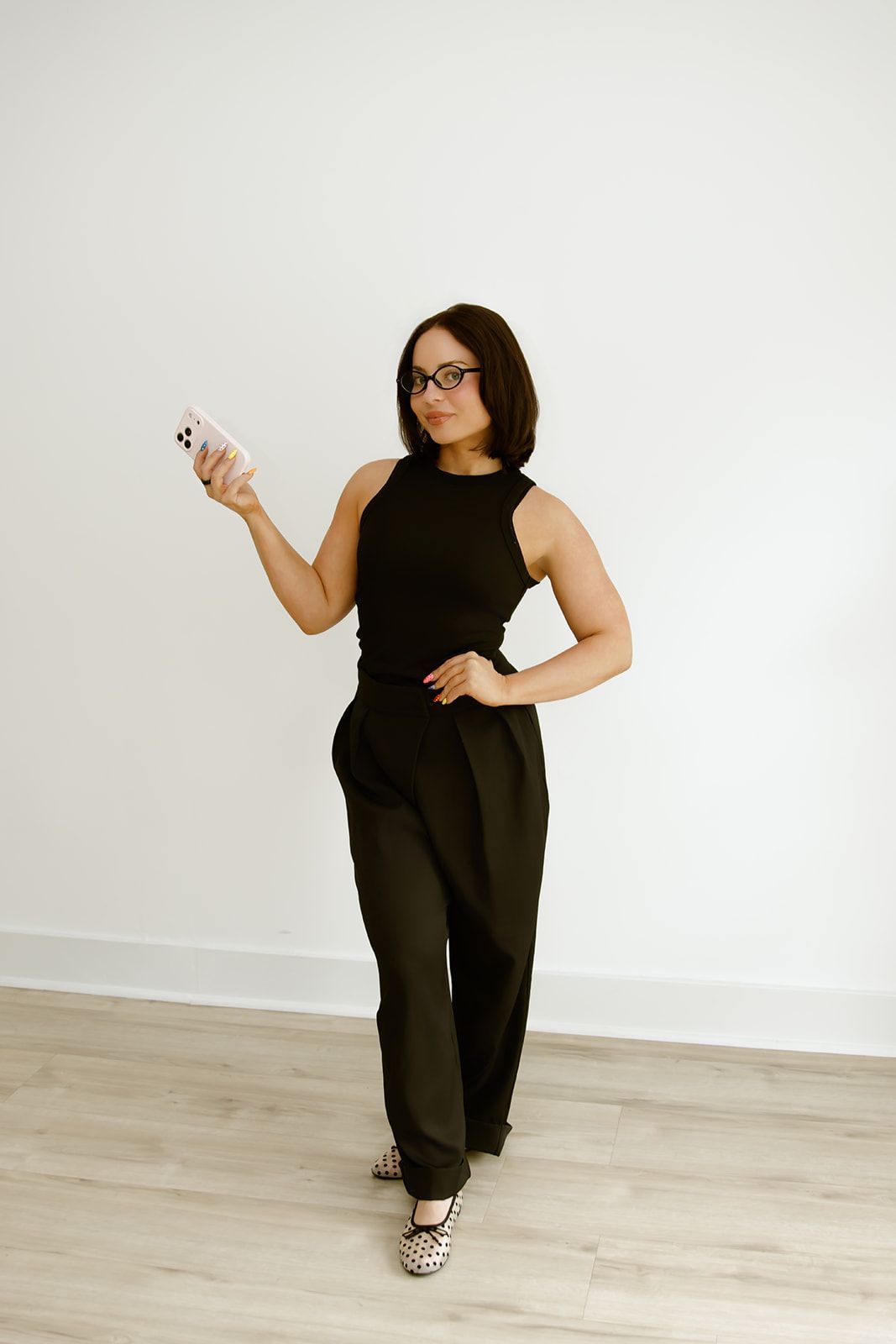 Person in a black sleeveless outfit posing against a white wall, holding a phone and standing on a wood floor.