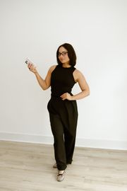 Person in a black sleeveless outfit posing against a white wall, holding a phone and standing on a wood floor.