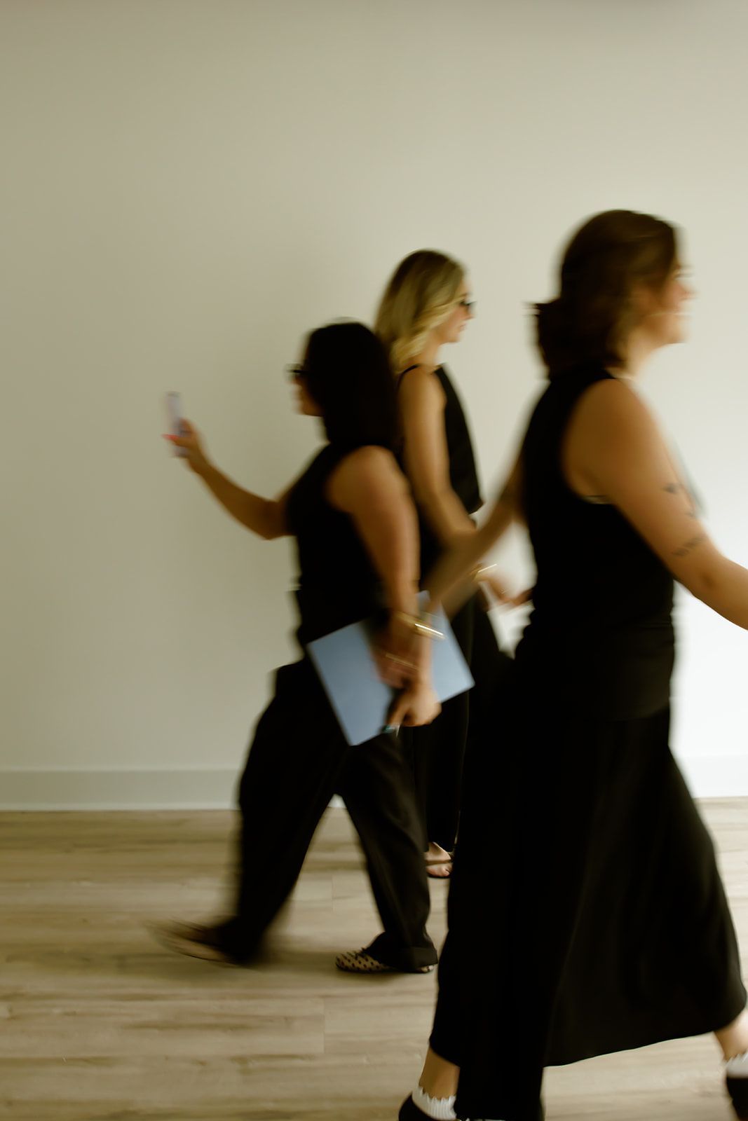 Three people walk past a blank wall in a dance studio, one holding a paper