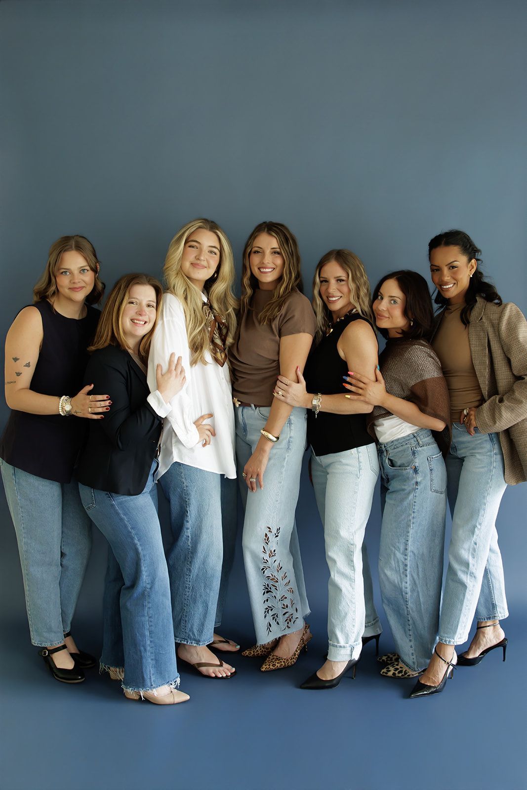 Six people standing together, smiling, in casual jeans and tops against a blue backdrop.