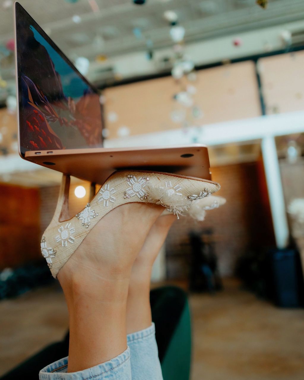 Hand holding a rose-gold laptop with a sparkly silver high heel balancing on top