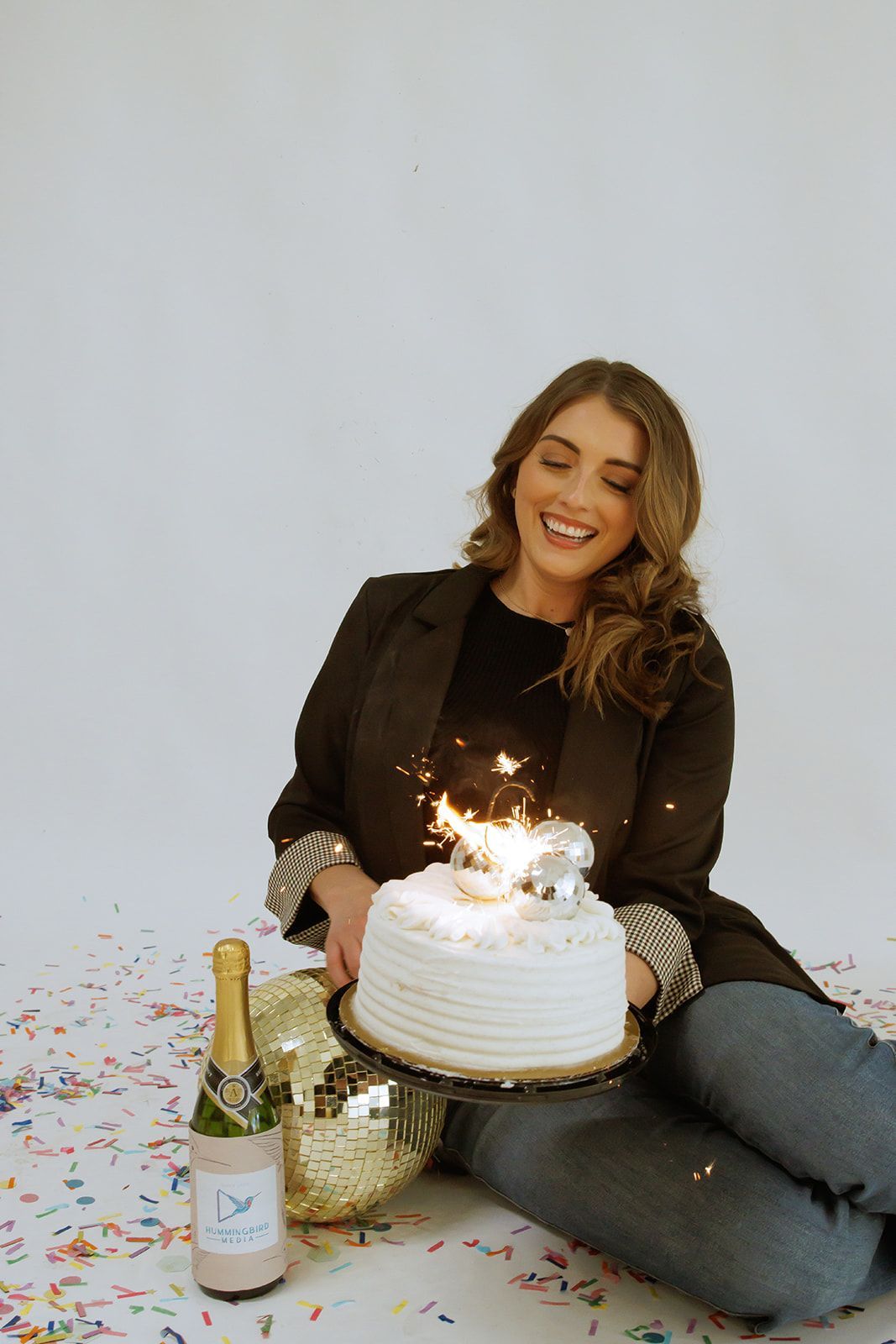 Woman smiling beside a lit birthday cake, champagne bottle, and gifts on a white backdrop