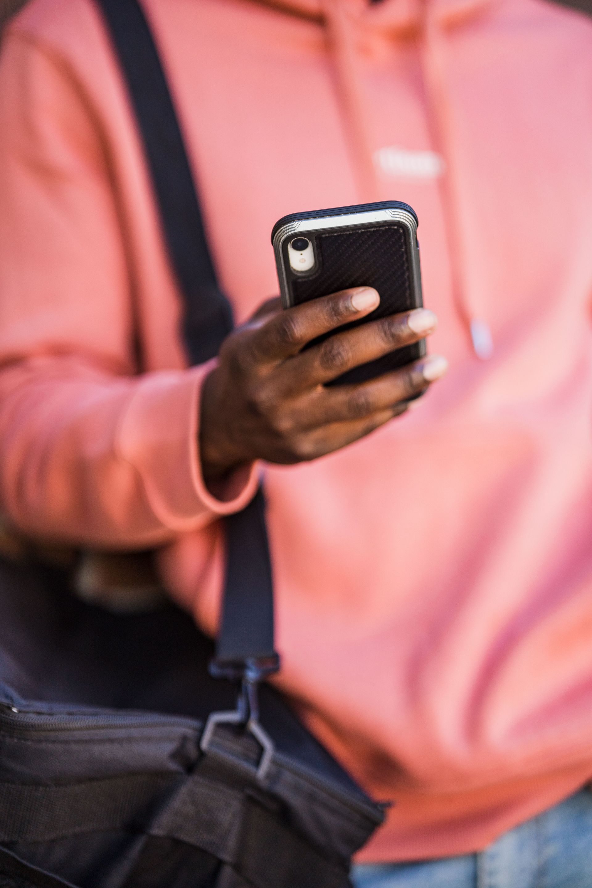 Person in a pink sweatshirt holding a smartphone with a black crossbody bag strap visible