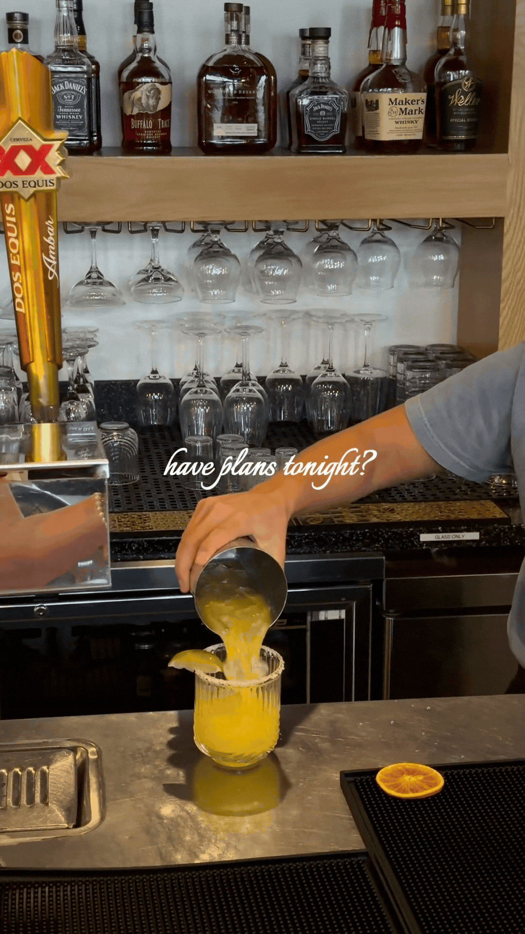 Bartender pours a yellow cocktail into a glass at a bar with liquor bottles and glassware behind it.