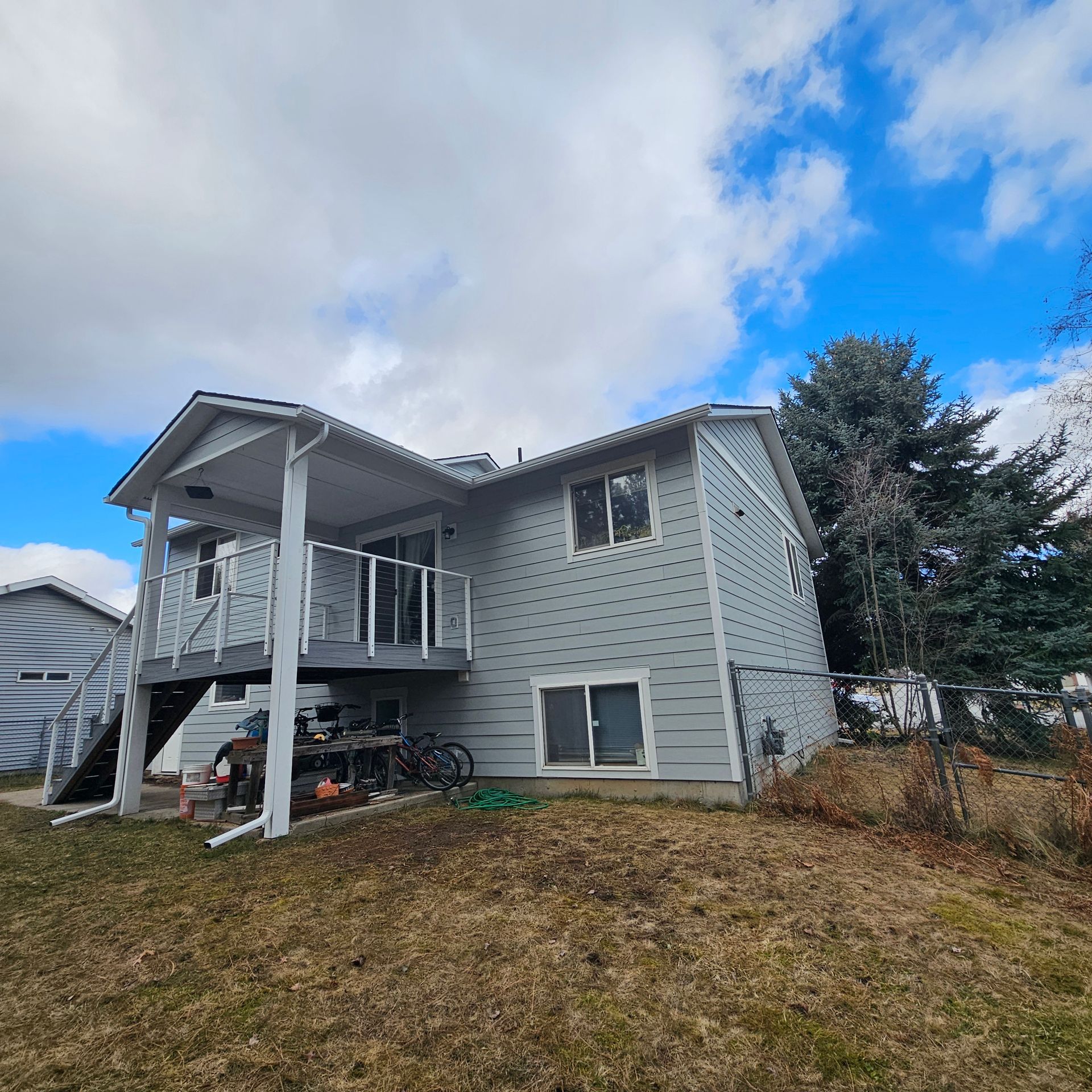 Gray house with a deck and stairs, set on a grassy hill under a partly cloudy blue sky.