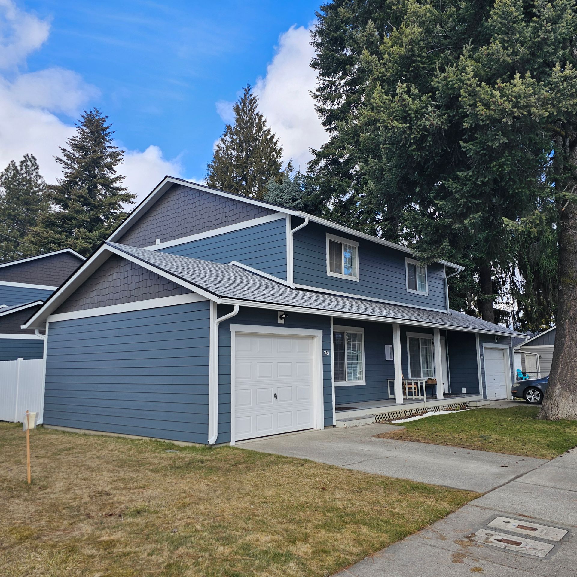 Two-story blue house with white trim and a white garage door, trees in the background, gray roof.