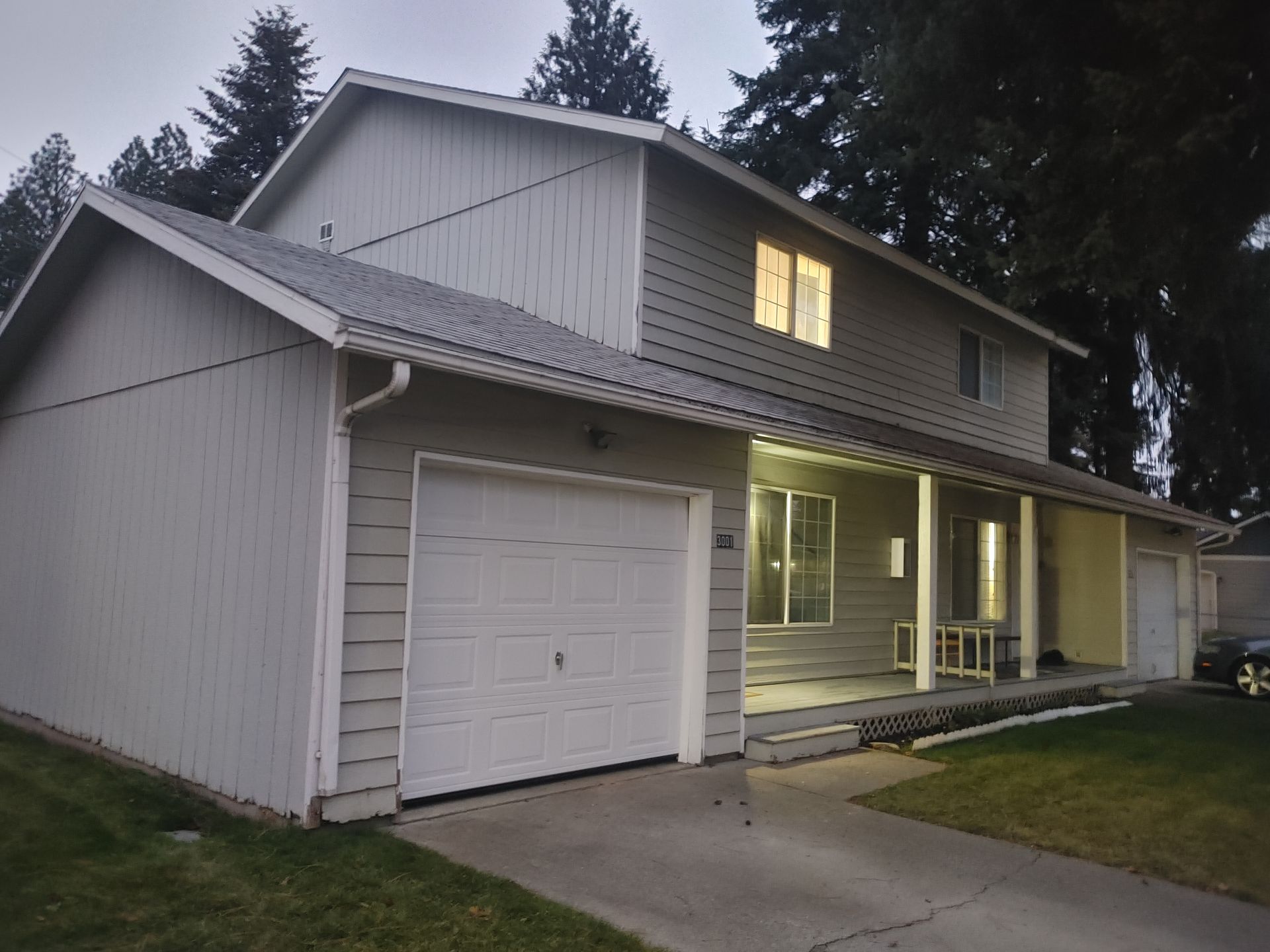 Two-story house with a white garage. Porch with lights on, grass lawn and dark trees in background.