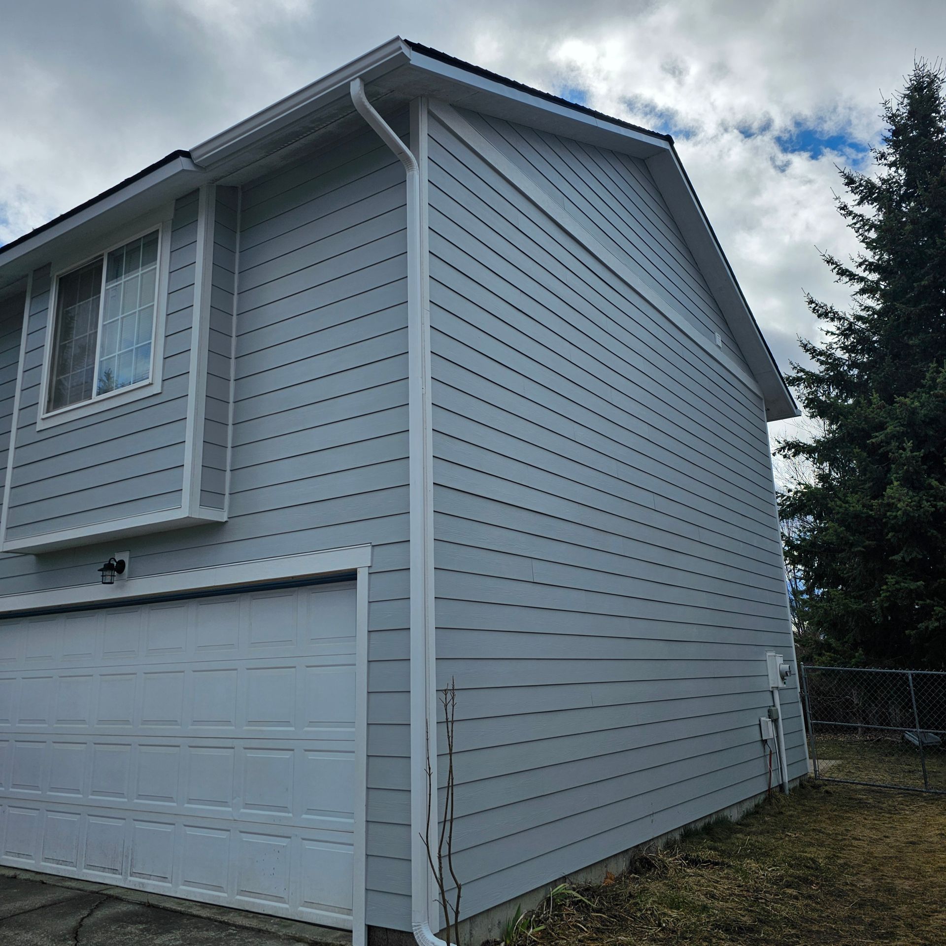 Light blue two-story house with white trim, garage door, and a partially visible tree on a cloudy day.