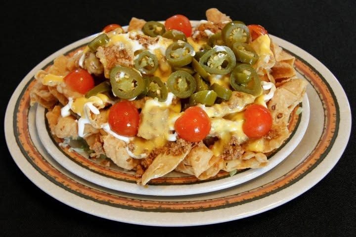 Loaded plate of nachos topped with melted cheese, jalapeños, cherry tomatoes, and olives