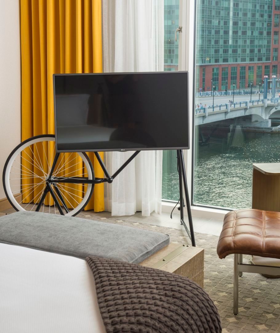 A flat-screen TV mounted on a bicycle frame stand in a hotel room with a bridge view.