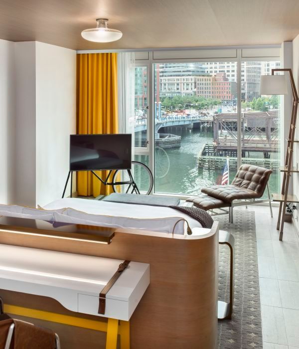 Modern hotel room overlooking a harbor. Features a bed, desk, and a lounge chair, with a bright yellow curtain.