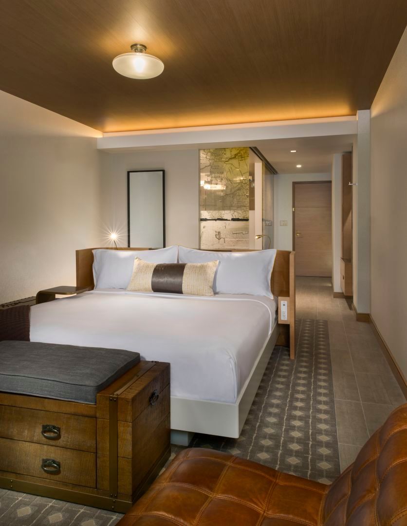 Modern hotel room with a bed, bench, and accent chair. Neutral colors with wood accents.