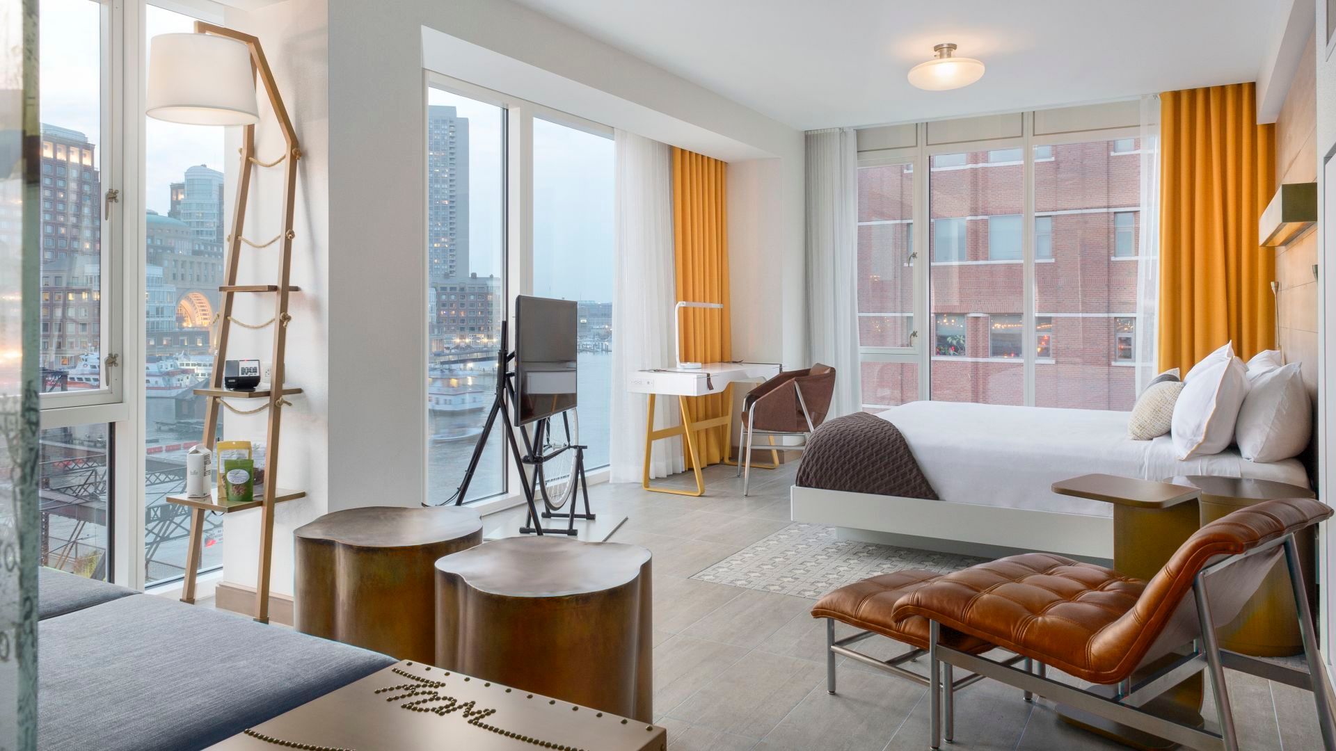 Modern hotel room with city views, featuring a bed, seating area, desk, and large windows.